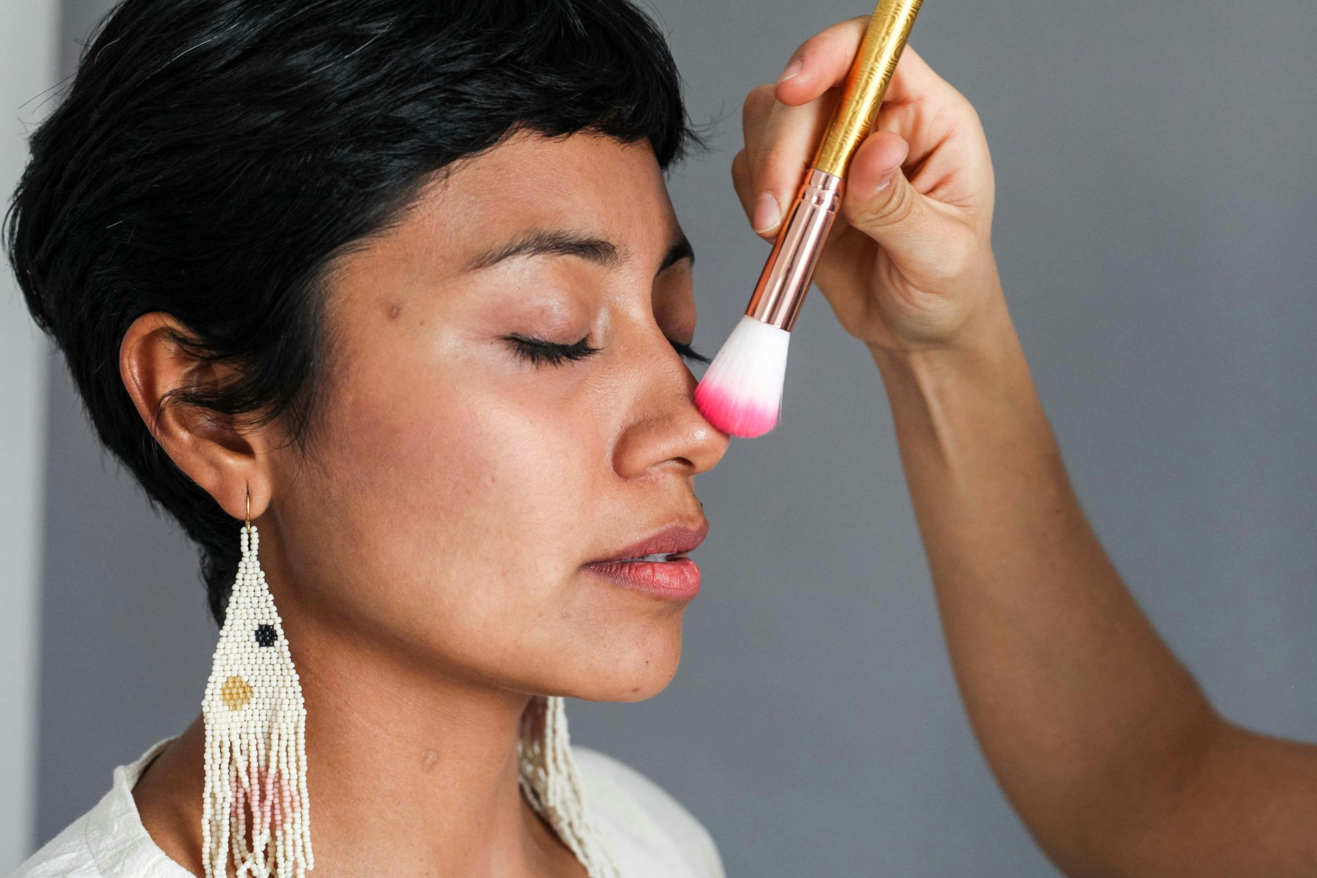 A woman with closed eyes applying makeup with a brush on her nose, showcasing beauty routines.