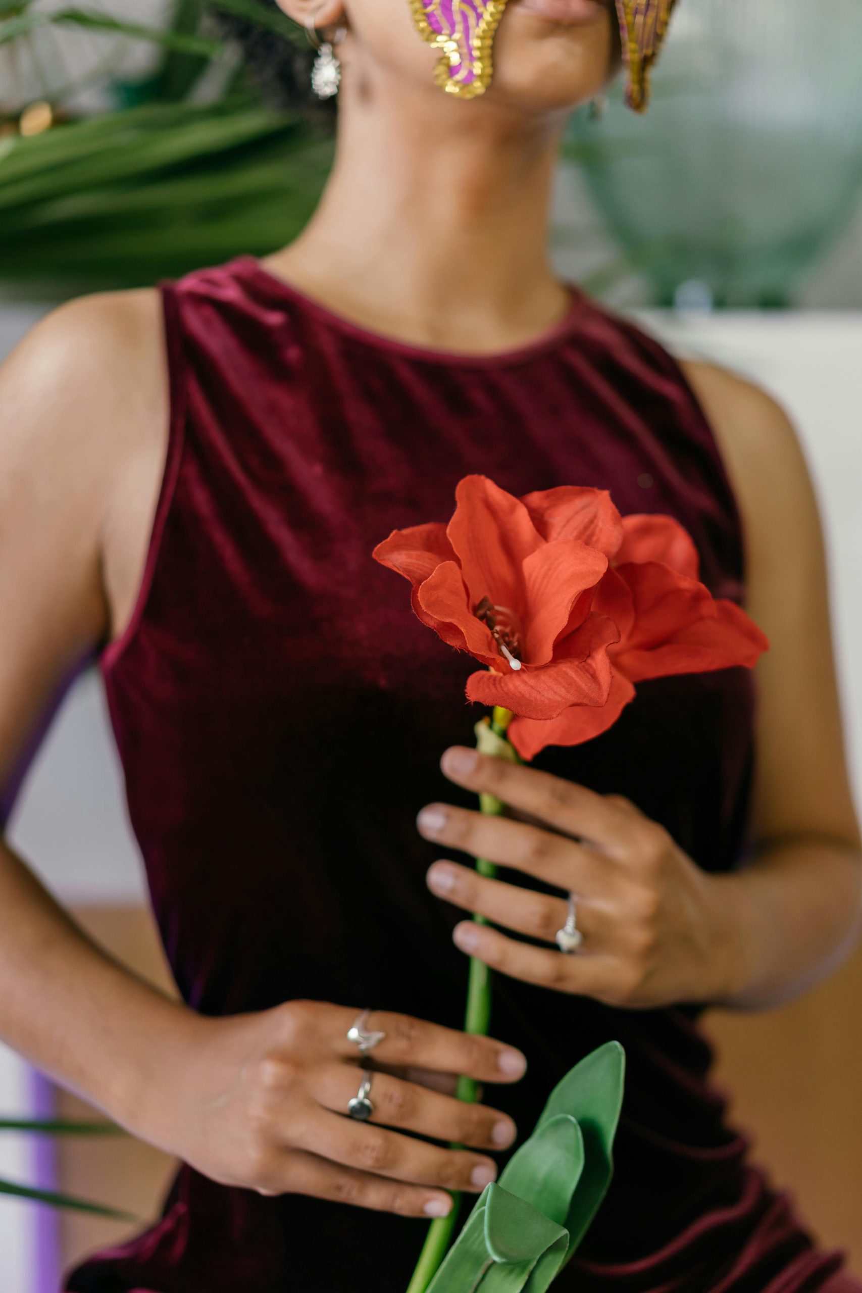 An elegant woman in a velvet dress holds a vibrant red flower, wearing a decorative mask.
